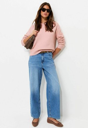 Mujer con gafas de sol negras, suéter rosa claro, vaqueros anchos azules, zapatos planos marrones y llevando un bolso marrón al hombro sobre un fondo blanco.