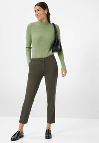 Olivgrüner gerippter Pullover mit hohem Kragen, kombiniert mit olivgrünen geraden Hosen und schwarzen Loafern; hält eine kleine schwarze Handtasche.
