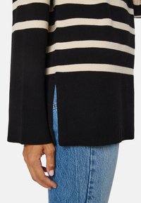 Pullover a strisce nere e beige con polsini a costine e aperture laterali, realizzato in un morbido materiale a maglia. Dettaglio ravvicinato della mano e della texture.