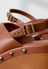 Sandali platform in pelle marrone con cinturini alla caviglia regolabili, dotati di fibbie in ottone e borchie in tonalità rame lungo la suola in legno.
