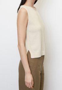 Top senza maniche color crema con scollatura tonda, texture a maglia, spacchi laterali e silhouette rilassata, abbinato a pantaloni fitted marroni.