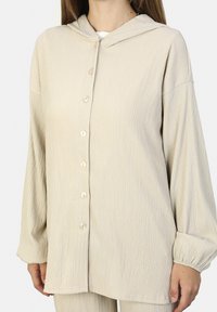 Beige, strukturierte Button-up-Bluse mit Kapuze, langen Ärmeln und Knopfverschluss. Sie verfügt über gerippten Stoff und elastische Bündchen.