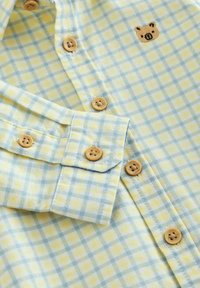 Hele kollase ja sinise ruuduline särk, millel on puidust nööbid ja rinnal tikitud seaprojekt. Puuvillane kangas, pehme tekstuuriga.