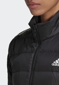 Chaqueta de plumas negra con un alto cuello, cierre de cremallera, patrón de acolchado horizontal y logo blanco de Adidas en el lado izquierdo del pecho.