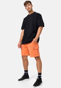 T-shirt en coton noir oversize associé à un short cargo orange avec poches latérales et détail de logo. Chaussures de sport noires et chaussettes rayées.