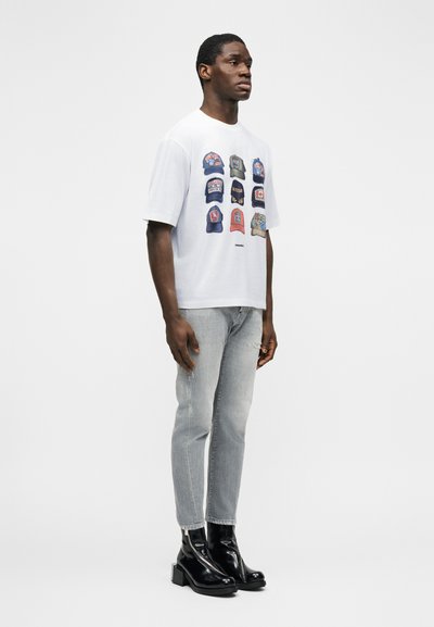 Άνδρας που στέκεται φορώντας λευκό T-shirt με εννέα πολύχρωμα σχέδια καπέλων, ανοιχτό γκρι τζιν και λαμπερά μαύρα μποτάκια αστραγάλου σε απλό φόντο.