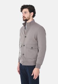 Giacca grigia chiara con colletto alto, chiusura a bottoni e due tasche frontali. Tessuto liscio e polsini a costine. Design semplice e moderno.