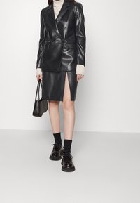 Kvinna i svart fodrad läder dubbelknäppt kavaj och kjol med hög slits, beige polotröja, svarta strumpor, kraftiga skor och handväska.