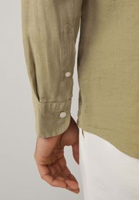 Camicia di lino verde oliva chiaro con maniche e polsini a punta, dotata di due bottoni e una texture sottile. Mano parzialmente visibile.