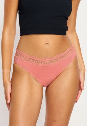 Femme portant des culottes roses bordées de dentelle et un top court noir, posant avec les bras détendus sur les côtés sur un fond uni.