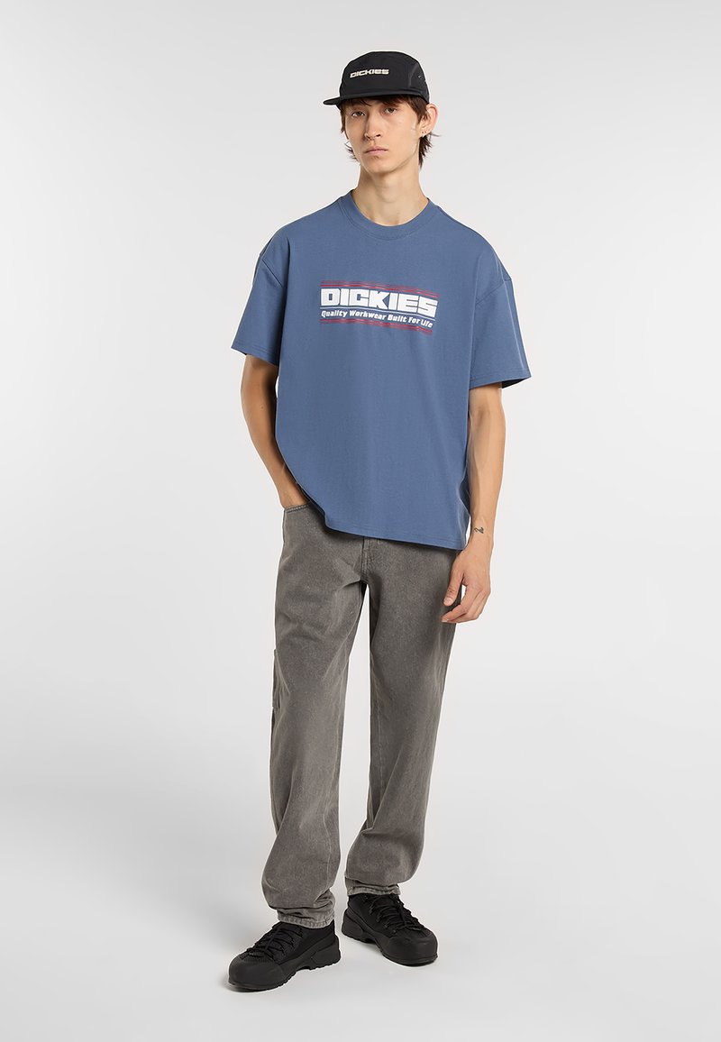 Camiseta de algodón azul con un logo gráfico audaz en blanco y rojo, combinada con pantalones gris claro y zapatillas negras. La camiseta tiene un ajuste holgado.