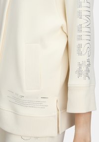 Sweatshirt crème clair avec des accents côtelés, une poche latérale et des détails en strass avec la phrase "LA JOIE D'ÊTRE" près de l'ourlet.