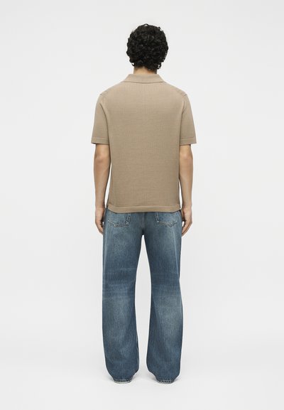 Άνδρας με σγουρά μαλλιά που φοράει μπεζ κοντομάνικο πόλο πουκάμισο και φαρδιά μπλε τζιν παντελόνια, στέκεται με την πλάτη προς απλό φόντο.