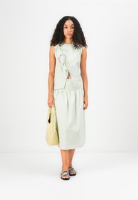 Top senza maniche verde chiaro con lacci laterali, abbinato a una gonna coordinata. Gli accessori includono una borsa a spalla beige e sandali con motivo.