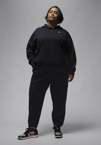 Felpa nera con cappuccio, tasca frontale e logo, abbinata a pantaloni della tuta neri coordinati. Indossata con sneakers nere e rosse.