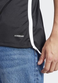 Maglietta sportiva nera con accenti bianchi ai lati, realizzata in tessuto liscio. Logo AEROREADY stampato in bianco vicino all'orlo inferiore. Abbinata a jeans blu.
