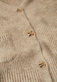 Cardigan en maille côtelée beige avec trois boutons brillants en écaille et une petite poche avant visible sur un tissu texturé.