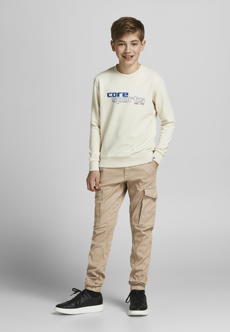 Svijetlosmeđi dukserica s grafikom "core sports", u kombinaciji sa svijetlo smešnim cargo pantalonama s bočnim džepovima. Crne patike upotpunjuju izgled.