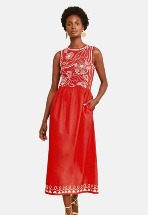 Frau in ärmellosem rotem Kleid mit weißer Blumenstickerei am Oberteil und Saum, steht mit einer Hand in der Tasche.
