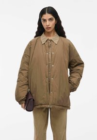 Kvinna med långt mörkt hår som bär en oversized brun jacka med tryckknappar, matchande byxor, och håller en mörkbrun kuvertväska i ena handen.