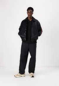 Jaqueta bomber preta com uma superfície texturizada, combinada com um hoodie preto e calças de ganga escuras e largas. Ténis beges e brancos finalizam o visual.