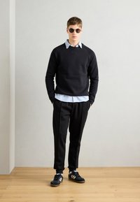 Black sweatshirt over a light blue striped shirt, paired with black trousers and black sneakers featuring white stripes. Round sunglasses complete the look.