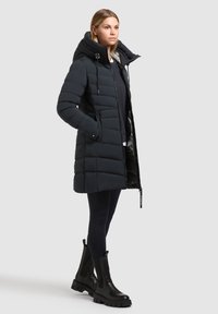 Casacondur de tip puffer, de culoare albastru închis, cu guler înalt, design matlasat și buzunare frontale, asortat cu cizme negre până la gleznă și colanți navy.