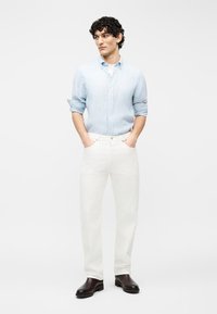 Άνδρας με κατσαρά μαλλιά, που φοράει γαλάζιο πουκάμισο με κουμπιά, λευκό παντελόνι και σκούρα καφέ μποτάκια, στέκεται με τα χέρια στις τσέπες.