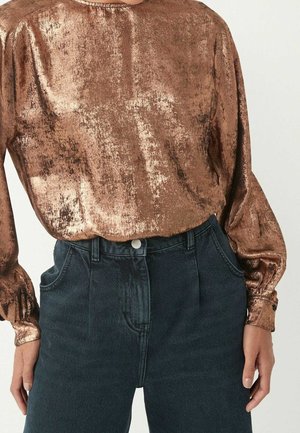 Kobieta ubrana w błyszczącą, miedzianą, długą bluzkę w rękawach wsuniętą w ciemnoniebieskie, wysokim stanem jeansy z przednimi kieszeniami.