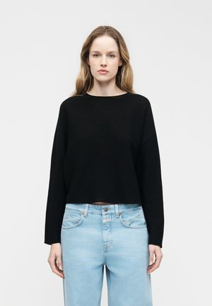 Młoda kobieta o jasnobrązowych włosach, ubrana w czarną bluzkę z długim rękawem i jasnoniebieskie dżinsy z wysokim stanem, stoi na tle jednolitego szarego tła.