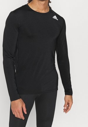 Homme portant un t-shirt noir ajusté à manches longues Adidas avec logo sur l'épaule gauche, debout, bras relâchés le long du corps.