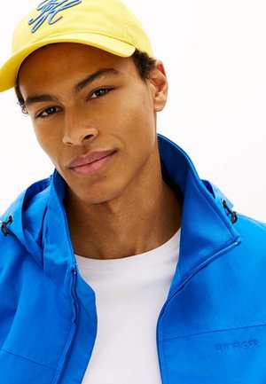 Giovane uomo con cappellino giallo, giacca blu brillante con cerniera e camicia bianca, volto verso lo spettatore con espressione neutra su sfondo bianco.