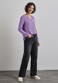 Blusa larga de manga larga en color púrpura con escote en V y detalles fruncidos, combinada con pantalones vaqueros de pierna ancha negros y zapatillas blancas sobre un suelo gris texturizado.