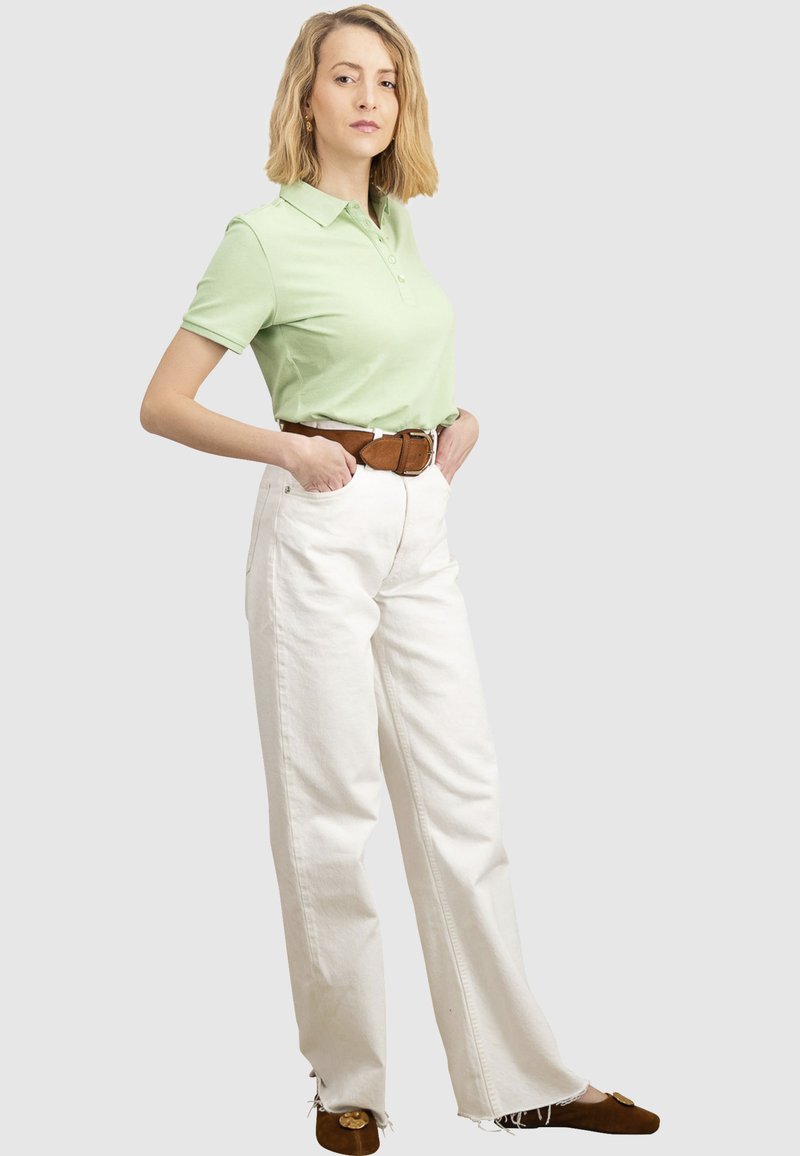 Žena stoji s rukama u džepovima, nosi svijetlo zelenu polo majicu, bijele široke hlače, smeđi remen i smeđe ravne cipele.