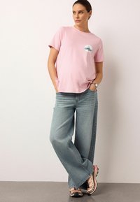Roze T-shirt met korte mouwen en een ronde zakprint, gecombineerd met lichtblauwe jeans met wijde pijpen en neutrale sneakers met accenten.