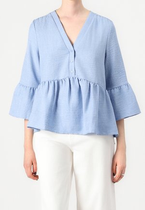 Femme portant un chemisier texturé bleu clair avec trois boutons, col en V, manches évasées, taille peplum et pantalon blanc.