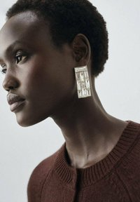De grandes boucles d'oreilles géométriques en argent avec des découpes rectangulaires et carrées transparentes, portées par un mannequin aux cheveux courts et foncés, sur un fond clair.