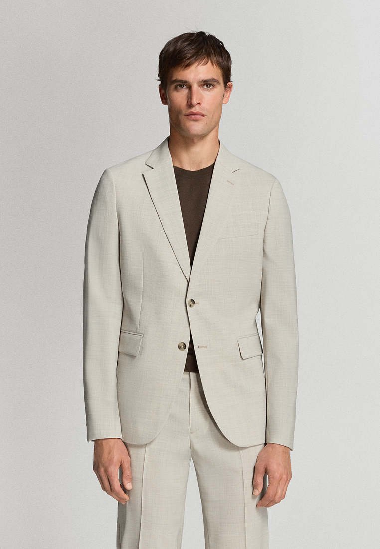 Homme portant une veste de costume beige clair à deux boutons et un pantalon assorti sur un t-shirt col rond marron foncé, debout devant un fond clair uni.