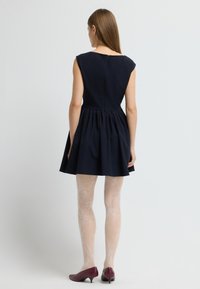 Une femme se tient de dos, portant une robe noire sans manches, des collants à motifs blancs et des chaussures à petit talon bordeaux, le tout sur un fond uni.