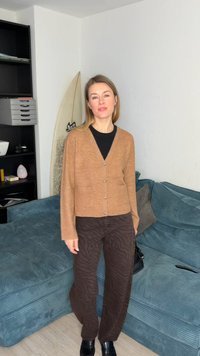 Femme debout dans une pièce portant un cardigan beige, un pantalon foncé à motifs et des chaussures noires, à côté d'un canapé turquoise et d'une planche de surf.