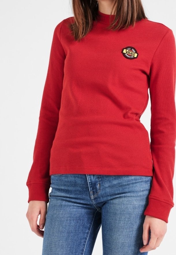 Femme portant une chemise Vans à manches longues rouges avec un petit logo sur la poitrine et un jean en denim bleu, se tenant devant un fond blanc.