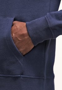 Mörkhyad hand insatt i framfickan på en marinblå sweatshirt med ribbad manschett och synliga sömnadsdetaljer.