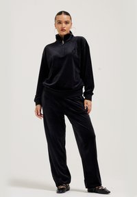 Vrouw in een zwart velours trainingspak met een ritskraag, grote oorringen en zwarte balletschoenen, staande tegen een effen achtergrond.