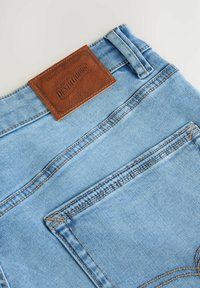 Blugi din denim albastru deschis, cu un etichetă texturată din piele maro la talie, având cusături contrastante și buzunare pe spate.
