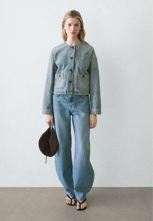 Donna con giacca di denim grigia abbottonata, jeans blu larghi, infradito nere, che tiene una borsa a sacchetto in suede marrone contro una parete liscia.