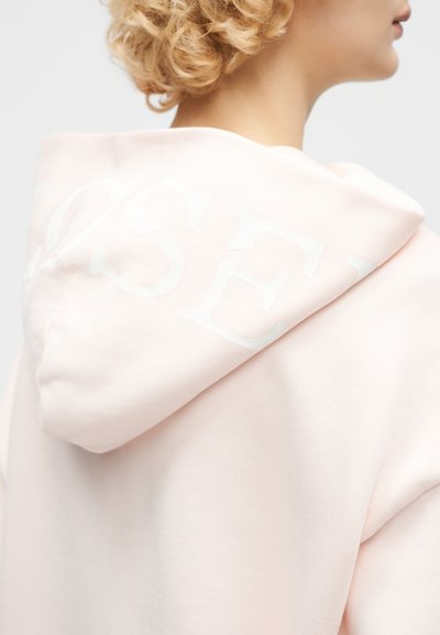 Κοκκαλάκι ξανθιάς με σγουρά μαλλιά, φορούσα ένα ανοιχτό ροζ φούτερ με μια αχνή λευκή λογότυπο στη κουκούλα, εμφανίζεται από πίσω και πλευρά.