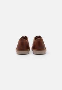 Chaussures en cuir marron avec une texture lisse, des coutures orange contrastantes et une semelle en caoutchouc de couleur claire, vues de l'arrière.