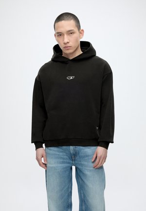 Νεαρός άνδρας με κοντά μαλλιά που φοράει μαύρο φούτερ με μια μικρή λευκή λογότυπο, συνδυασμένο με ανοιχτό μπλε τζιν, στέκεται μπροστά σε ένα απλό λευκό φόντο.