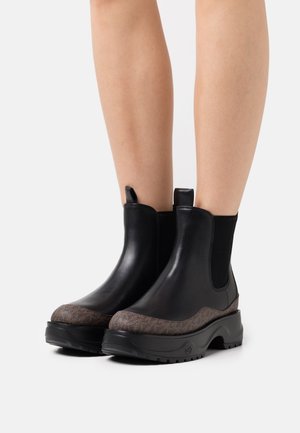 Bottes basses en cuir noir avec panneaux latéraux élastiques, accent brun texturé sur le bout, et semelle en caoutchouc noir robuste.