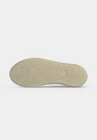 Semelle de sneaker beige avec motif antidérapant texturé et petit logo rectangulaire près du talon sur fond blanc.